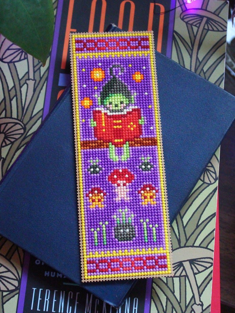 May include: A handmade bookmark with a purple background and a stitched design. The design features a green creature reading a red book, with mushrooms and stars. The bookmark has a yellow border and a black ribbon.