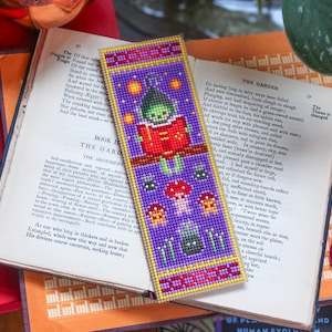 May include: A beaded bookmark with a fantasy theme. The purple bookmark depicts a green character reading a red book, with mushroom and star motifs. It has red and yellow borders. The bookmark is placed on an open book, with text visible.