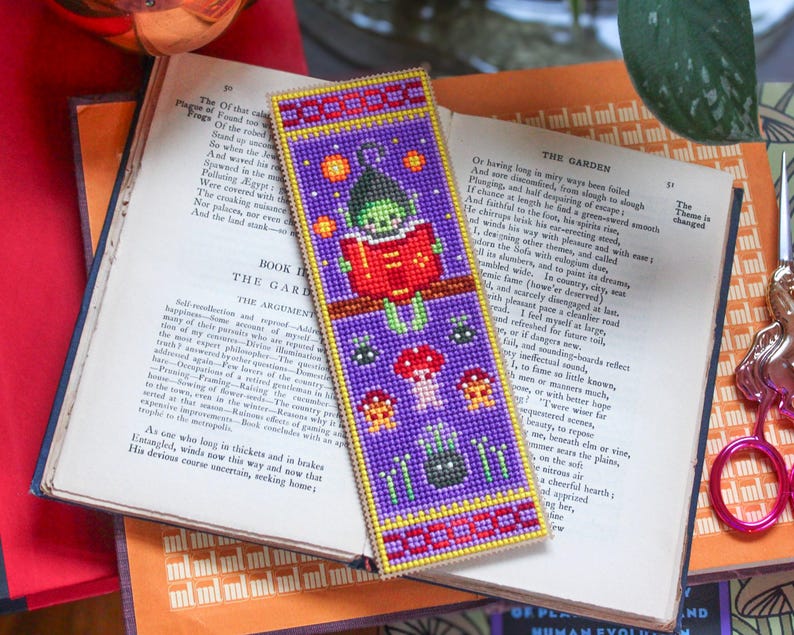 May include: A beaded bookmark with a fantasy theme. The purple bookmark depicts a green character reading a red book, with mushroom and star motifs. It has red and yellow borders. The bookmark is placed on an open book, with text visible.