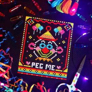 Könnte beinhalten: Ein farbenfrohes Kreuzstichmuster eines Clowns mit dem Text "PEG ME" auf schwarzem Hintergrund. Das Muster ist von einem Regenbogenrand umgeben.