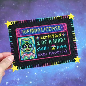 Op de afbeelding: Een kruissteek "Weirdo License" met een cartoon kat illustratie. De zwarte achtergrond heeft tekst en een kleurrijke rand. Gele ster accenten zijn bevestigd aan de hoeken. De licentie luidt "certified 1 of a kind!" en "skill: probing".