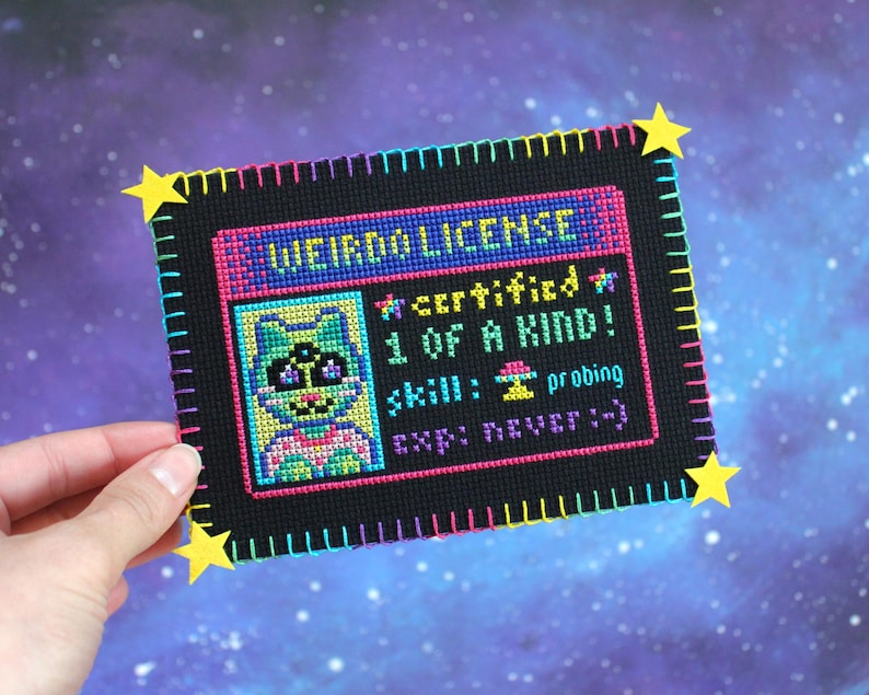 Op de afbeelding: Een kruissteek "Weirdo License" met een cartoon kat illustratie. De zwarte achtergrond heeft tekst en een kleurrijke rand. Gele ster accenten zijn bevestigd aan de hoeken. De licentie luidt "certified 1 of a kind!" en "skill: probing".