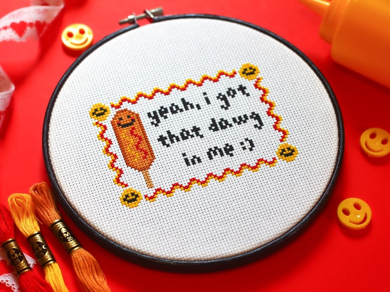 May include: A white embroidery hoop with a black frame, featuring a cross-stitch design of a smiling corn dog with the text "yeah, i got that dawg in me :)".