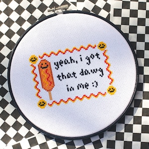 May include: A white embroidery hoop with a red and yellow border. The hoop features a cross-stitch design of a corn dog with the text "yeah, i got that dawg in me :)"
