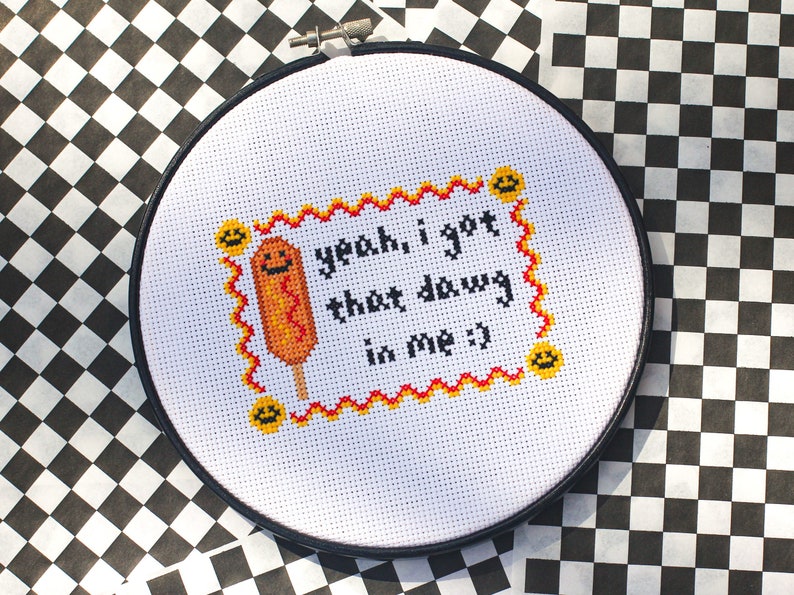May include: A white embroidery hoop with a red and yellow border. The hoop features a cross-stitch design of a corn dog with the text "yeah, i got that dawg in me :)"