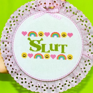 Puede incluir: Un bastidor de bordado de punto de cruz con un borde de encaje rosa y un lazo de cuadros. El dise&ntilde;o presenta la palabra "SLUT" en verde, rodeada de corazones, arco&iacute;ris y caras sonrientes. El fondo es blanco.