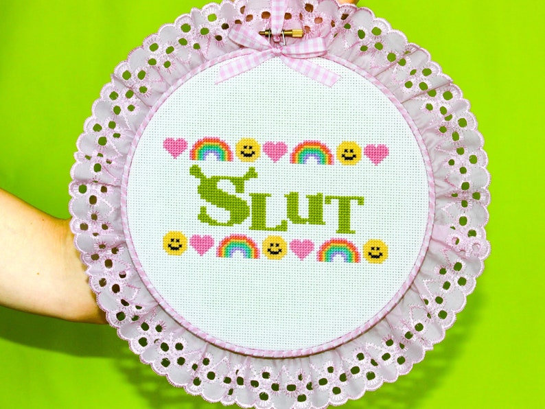 Puede incluir: Un bastidor de bordado de punto de cruz con un borde de encaje rosa y un lazo de cuadros. El dise&ntilde;o presenta la palabra "SLUT" en verde, rodeada de corazones, arco&iacute;ris y caras sonrientes. El fondo es blanco.