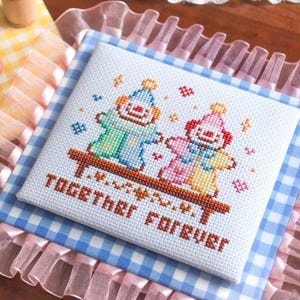 May include: A cross-stitch piece featuring two clown figures holding hands, with the words "TOGETHER FOREVER" stitched below. The artwork is set on a blue and white gingham pattern, surrounded by pink ruffled fabric.