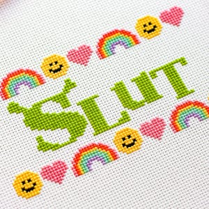 Puede incluir: Bordado de punto de cruz sobre tela blanca. La palabra "SLUT" est&aacute; bordada en verde, rodeada de arco&iacute;ris, caras sonrientes y corazones rosas. El dise&ntilde;o est&aacute; en un aro redondo.