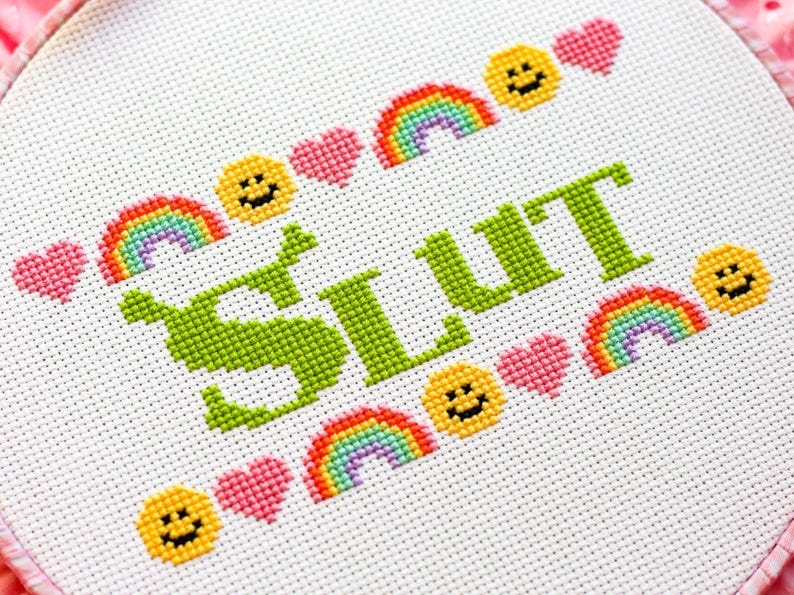 Puede incluir: Bordado de punto de cruz sobre tela blanca. La palabra "SLUT" est&aacute; bordada en verde, rodeada de arco&iacute;ris, caras sonrientes y corazones rosas. El dise&ntilde;o est&aacute; en un aro redondo.