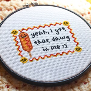 May include: A white embroidery hoop with a black frame, featuring a cross-stitched design of a yellow and orange corn dog with a smiling face. The text "yeah, i got that dawg in me :)" is stitched in black thread.