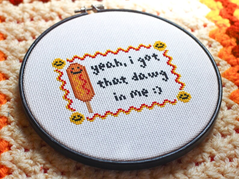 May include: A white embroidery hoop with a black frame, featuring a cross-stitched design of a yellow and orange corn dog with a smiling face. The text "yeah, i got that dawg in me :)" is stitched in black thread.