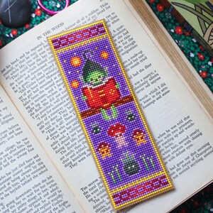 May include: A handcrafted bookmark with a purple backdrop and a witch motif. The bookmark depicts a witch reading, with stars, mushrooms, and a spider. The bookmark is placed on an open book.