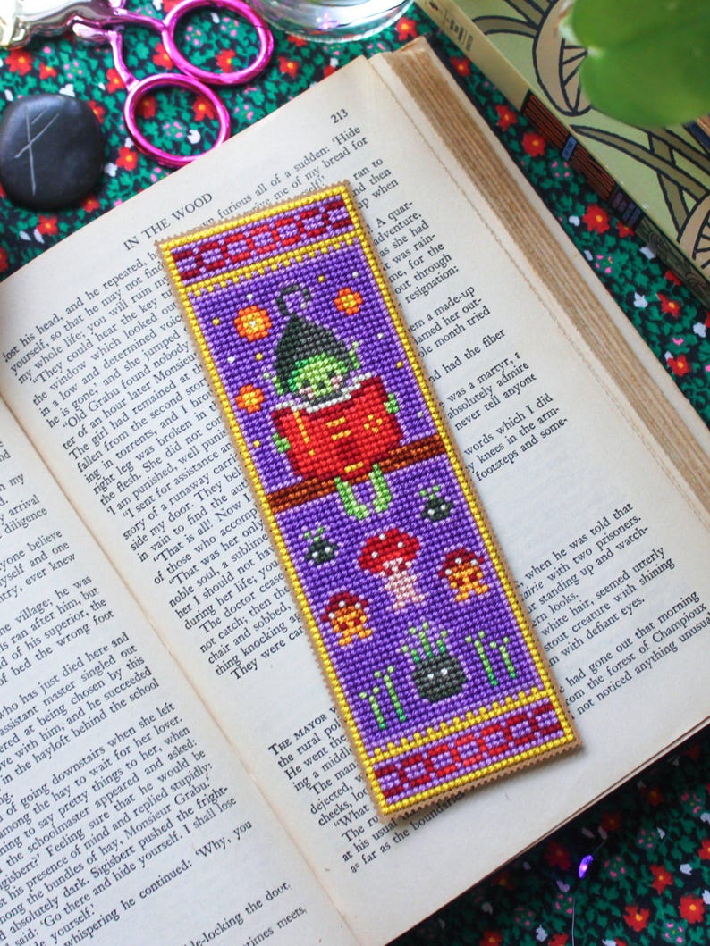 May include: A handcrafted bookmark with a purple backdrop and a witch motif. The bookmark depicts a witch reading, with stars, mushrooms, and a spider. The bookmark is placed on an open book.