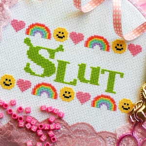 Puede incluir: Un bastidor de bordado de punto de cruz con la palabra "SLUT" en verde, rodeado de arco&iacute;ris, caras sonrientes y corazones rosas. Tambi&eacute;n se ven unas tijeras doradas con forma de unicornio y cuentas rosas con letras.