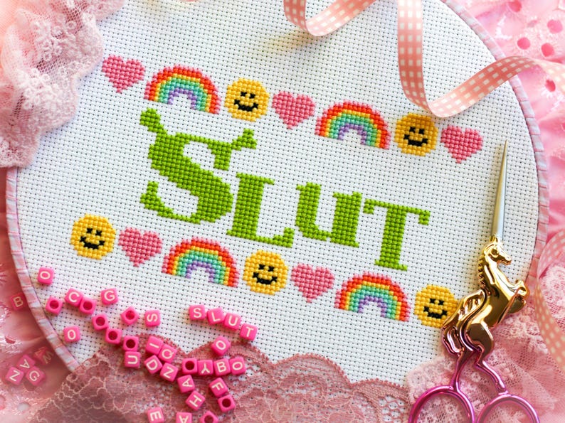 Puede incluir: Un bastidor de bordado de punto de cruz con la palabra "SLUT" en verde, rodeado de arco&iacute;ris, caras sonrientes y corazones rosas. Tambi&eacute;n se ven unas tijeras doradas con forma de unicornio y cuentas rosas con letras.