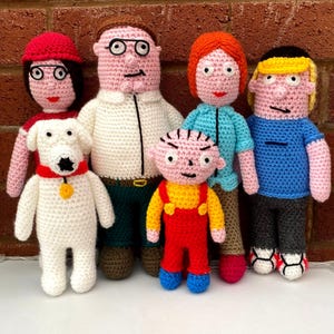 May include: A set of six crocheted dolls representing a family, including a white dog with a red collar, a man in a white shirt and green pants, a woman in a red hat, a woman in a blue shirt, a child in red overalls, and a child in a blue shirt.