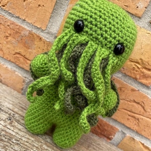 May include: A green crocheted plush toy featuring a rounded head with two black plastic eyes and multiple dangling tentacles covering the face area. The creature has a soft, textured yarn surface and two small arms extending from its sides.