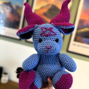 May include: A crocheted plush toy featuring a light blue body with dark magenta horns, ears, and feet. The creature has black plastic eyes, a stitched magenta nose, and a magenta pentagram embroidered on its forehead in a textured yarn finish.