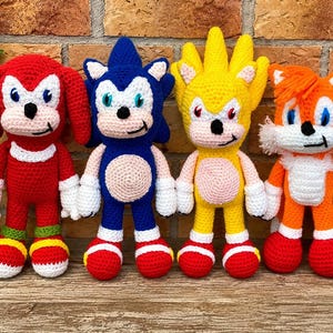 May include: Four handmade crochet dolls representing characters from the Sonic the Hedgehog series. The set includes a red Knuckles, a blue Sonic, a yellow Super Sonic, and an orange Tails, each featuring white accents, embroidered eyes, and stitched details.
