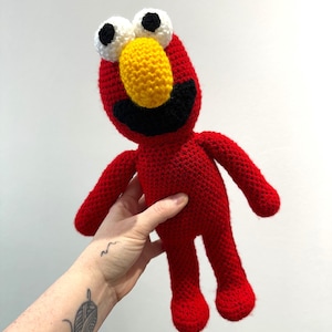 Może przedstawiać: Ręcznie robiona czerwona szydełkowa lalka z żółtym nosem, czarnymi ustami oraz białymi i czarnymi oczami. Lalka ma ręce i nogi i jest postacią z programu telewizyjnego dla dzieci.