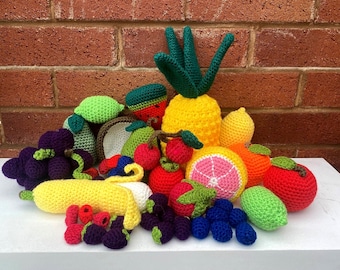 May include: A collection of various crocheted fruits and vegetables including a yellow pineapple, a peeled banana, purple grapes, red apples, a sliced grapefruit, a lime, a pear, and assorted berries in vibrant colors like red, blue, and green.