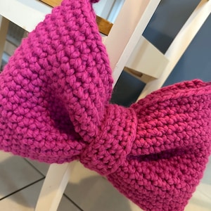 Pode incluir: Um grande laço de crochê rosa brilhante. O laço é feito com um fio grosso e tem uma superfície texturizada e irregular.