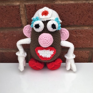 Puede incluir: Muñeca de ganchillo con forma de patata, cuerpo marrón, brazos y sombrero blancos y zapatos rojos. La muñeca tiene ojos grandes, nariz y orejas rosas y boca roja y blanca. El sombrero está adornado con una flor roja.