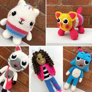 May include: A collection of handmade crochet dolls and figures. The dolls include a white cat with a rainbow striped belly, a yellow cat with pink accents, and other characters in various colors. The dolls are displayed against a brick background.