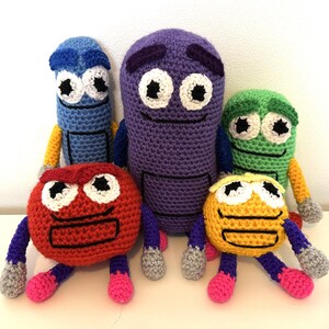 Puede incluir: Cinco juguetes de peluche de crochet con forma de lápices de colores. Los lápices son rojo, naranja, amarillo, verde y morado. Cada lápiz tiene una cara sonriente con ojos grandes y una boca simple.