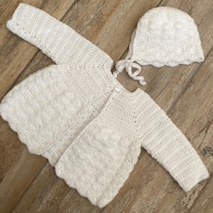 Puede incluir: Un cárdigan blanco de ganchillo para bebé y un gorro a juego. El cárdigan tiene cierre de botón y el gorro tiene un lazo.