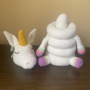 May include: A white crocheted unicorn toy with a yellow horn and a white crocheted stacking ring toy with purple, pink, and green accents.