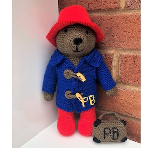 Puede incluir: Un oso de peluche de crochet que lleva un sombrero rojo, un abrigo azul y botas rojas. El oso lleva una maleta marrón con las iniciales "PB" en ella.