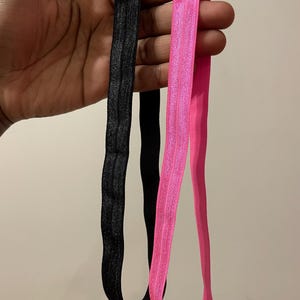 May include: Two hair ties, one black and one pink, are held in a hand. The hair ties are made of elastic material and are about 1/2 inch wide.