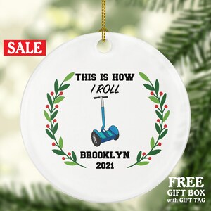 May include: White ceramic Christmas ornament with a green wreath and red berries. The ornament features a blue and white self-balancing scooter with the text "This is how I roll" and "Brooklyn 2021".