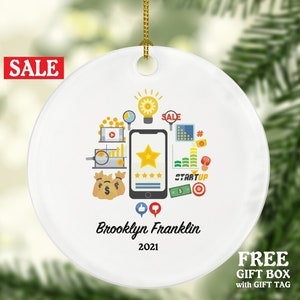 May include: White ceramic Christmas ornament with a colorful illustration of a smartphone with a five-star rating, a lightbulb, and other business-related icons. The text "Brooklyn Franklin 2021" is printed below the illustration.