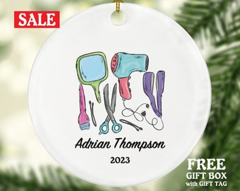 May include: White ceramic Christmas ornament with a colorful illustration of hairdressing tools, including a hairdryer, scissors, combs, and a mirror. The ornament is personalized with the name "Adrian Thompson" and the year "2023".