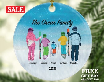 Peut inclure: Une boule de Noël ronde, bleue et blanche avec un fond enneigé. La boule représente une famille de cinq skieurs, avec le texte "The Oscar Family" et l'année "2021".
