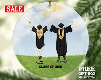 May include: A round ceramic ornament with a watercolor background of blue sky and green grass. Two figures in black graduation gowns with gold accents stand with their arms raised, holding diplomas. The text "Faith" and "Daniel" are below the figures, and "CLASS OF 2021" is below the names.