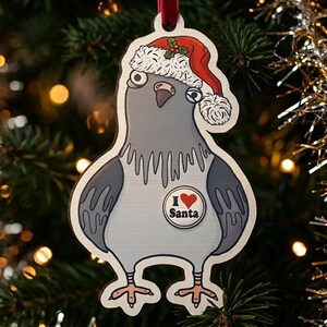 May include: A flat wooden Christmas tree ornament shaped like a pigeon wearing a red and white Santa hat with a green holly sprig. The pigeon has a white chest featuring a circular badge that reads I heart Santa with a red heart symbol.