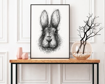 May include: A black and white line drawing of a rabbit's face in a black frame on a wooden table with a vase of branches and white stones.
