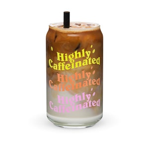 May include: A glass can with a straw filled with iced coffee. The can has the text "Highly Caffeinated" printed on it in yellow, orange, and pink.