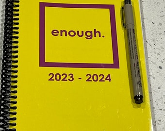 Pode incluir: Uma agenda amarela com uma encadernação em espiral preta. A capa tem um quadrado roxo com a palavra "enough" em letras brancas. A agenda é para os anos 2023-2024. Uma caneta bege está em cima da agenda.
