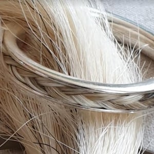 Könnte beinhalten: Ein silbernes Armband mit einem geflochtenen Band aus weißem Pferdeschwanz. Das Armband ist aus glattem, poliertem Metall gefertigt und hat ein schlichtes, elegantes Design.