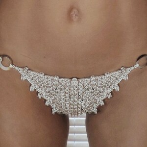 May include: A close-up of a crocheted bikini bottom with silver rings. The bikini bottom is made of a beige and white yarn.
