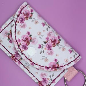 May include: A small rectangular pouch with a rounded flap featuring a white background with a pink and purple floral pattern. The pouch has dark red stitching along the edges, a white plastic snap closure, and a silver metal keyring attached.