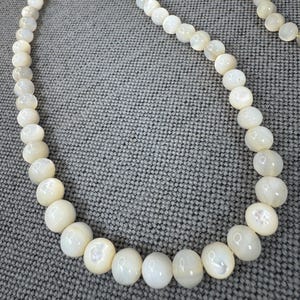 May include: A long, off-white beaded necklace. The round beads have a pearlescent sheen. The necklace is displayed on a grey textured surface.