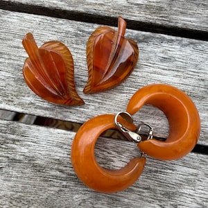Può includere: Un set di quattro gioielli color ambra composto da due clip a forma di foglia con dettagli venati intagliati e due orecchini a clip circolari a cerchio con meccanismi in metallo color argento sul retro.