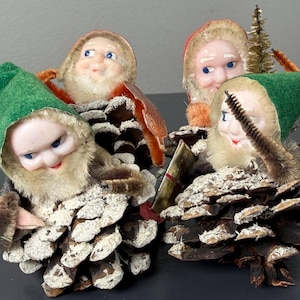 May include: Four vintage Christmas ornaments featuring elf-like figures with painted faces, green hats, and faux fur beards. The figures are attached to pine cones. One has a small bottle brush tree.