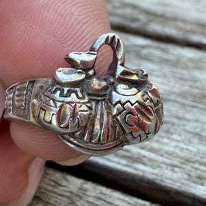 May include: A silver ring featuring a detailed, three-dimensional sculptural design on the band. The centrepiece consists of rounded, embossed shapes with intricate geometric patterns and a central loop, all finished in a polished metallic silver.